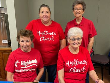 Four smiling women wearing red Highlands Church shirts indoors.