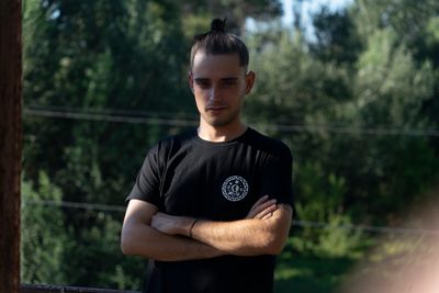 Young man with arms crossed, wearing a black t-shirt, standing outdoors with trees in the background.