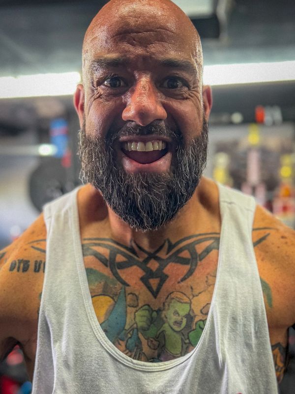 Smiling muscular man with a beard and colorful chest tattoos in a gym.