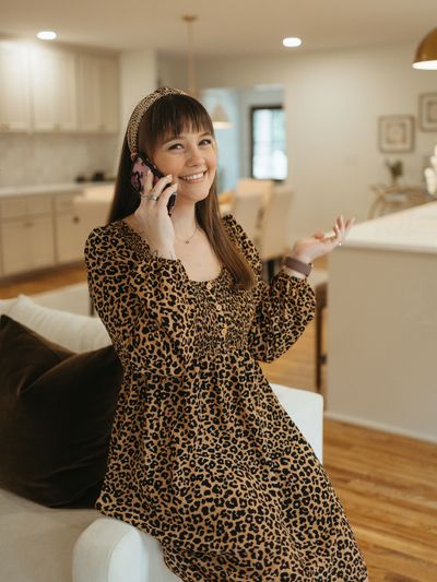 A smiling woman in a leopard print dress talks on the phone indoors.