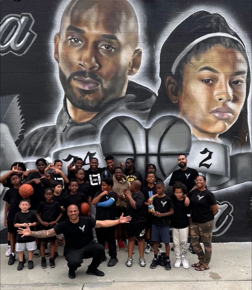 Group of kids and adults posing in front of a mural of Kobe Bryant and a young girl with basketball imagery.