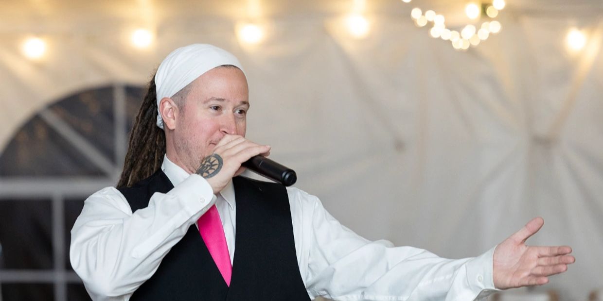 Man with dreadlocks and tattoo speaking into a microphone at an event.