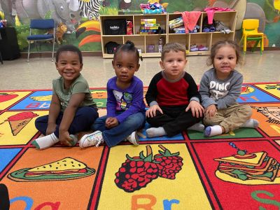 Four young children sitting on a colorful alphabet rug in a classroom.