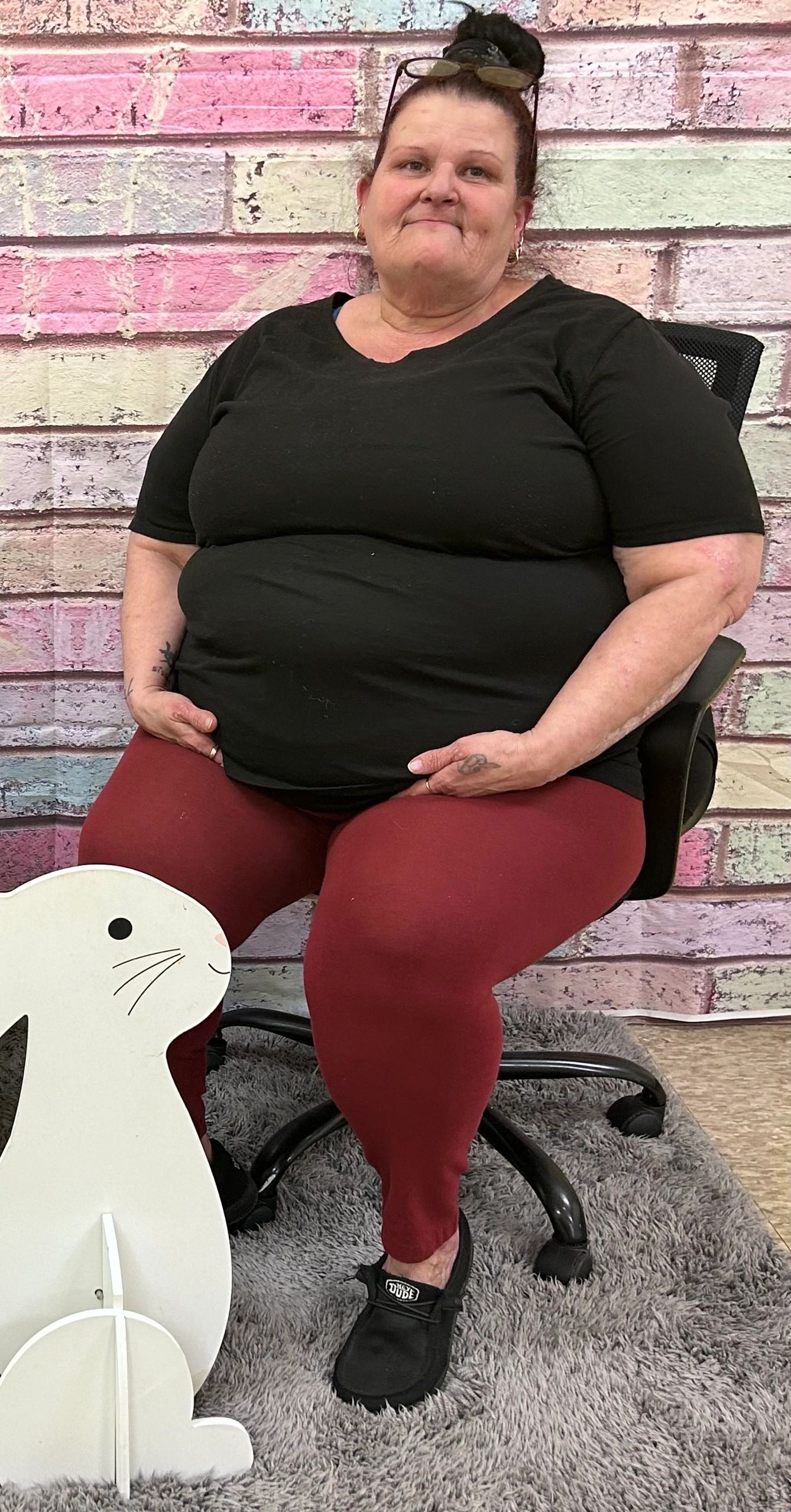 A woman in black and red sitting on a chair against a pastel brick wall.