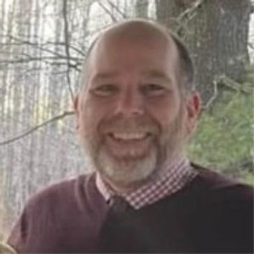 Smiling man in a maroon sweater standing outdoors with trees in the background.