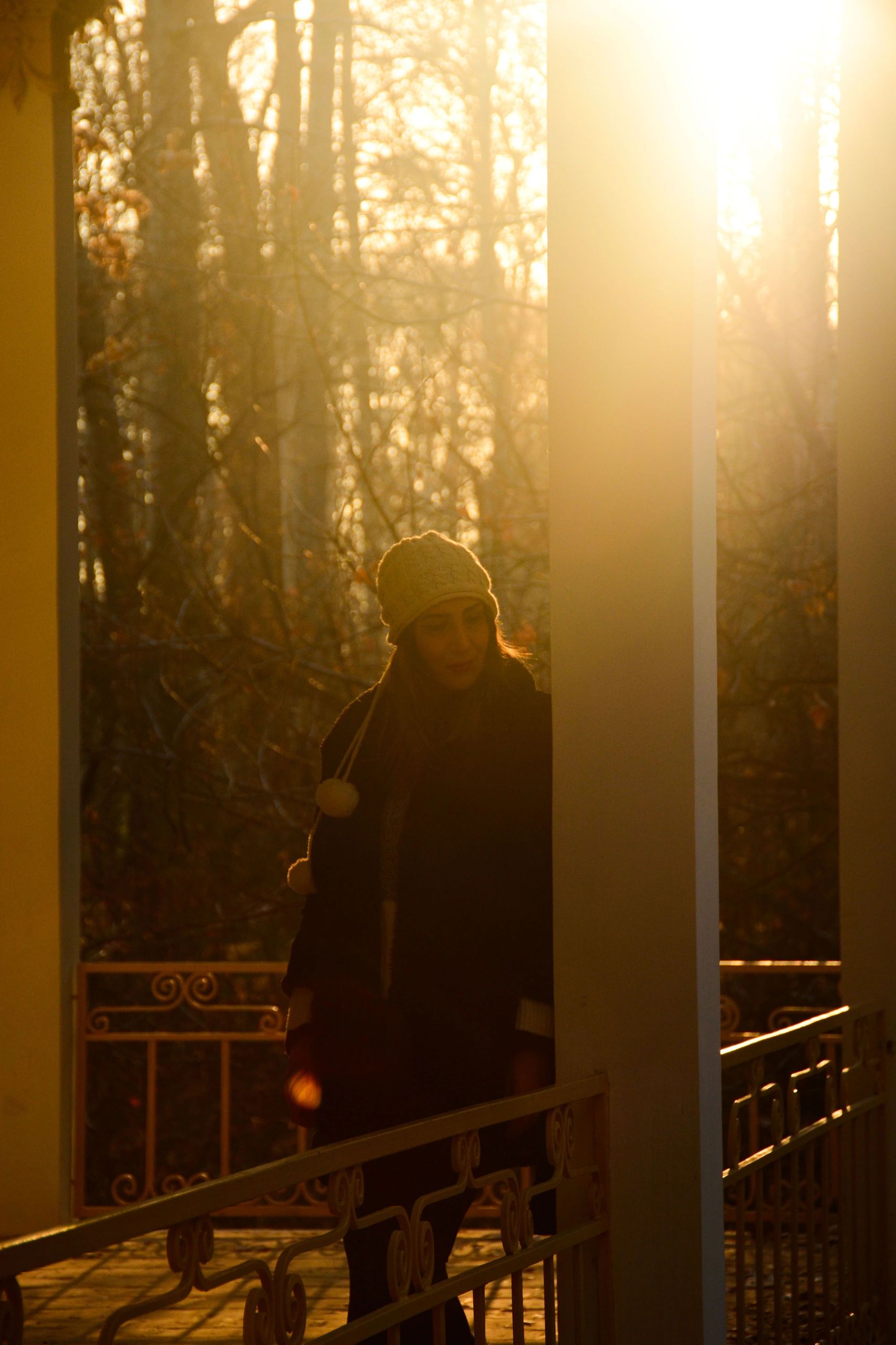 Woman in a cozy beanie leaning against a porch pillar in a sunlit park during golden hour. 