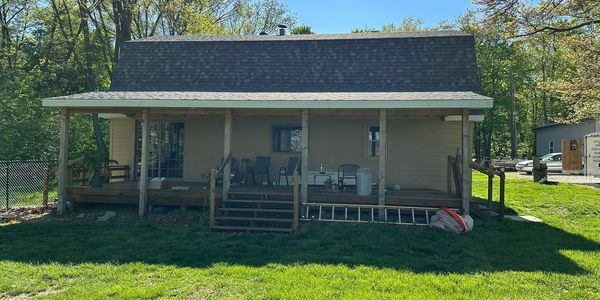 A modest house with a wooden porch surrounded by green grass and trees.