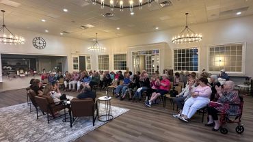 A group discussion in a well-lit community room with many seated attendees.