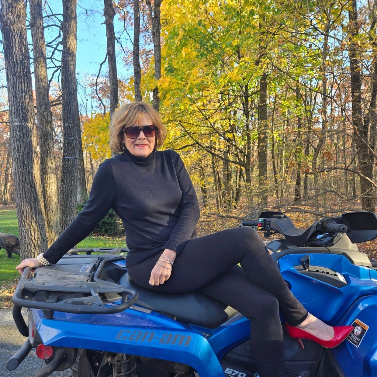 Woman in black outfit with red heels sitting on a blue Can-Am ATV in autumn woods.