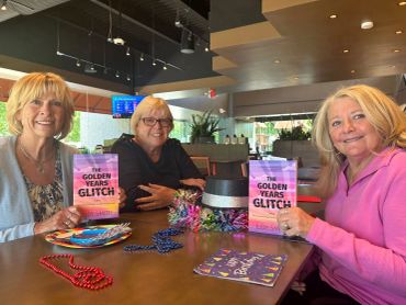 Three women proudly display copies of 'The Golden Years Glitch' at a cozy gathering spot.