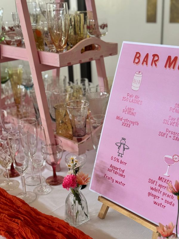 A pink bar menu on an easel with glassware and flowers on a table.