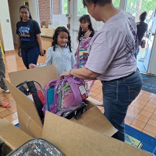 A woman distributes backpacks to children inside a building.