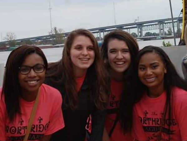 Six young women smiling outdoors near a waterfront with a bridge in the background.