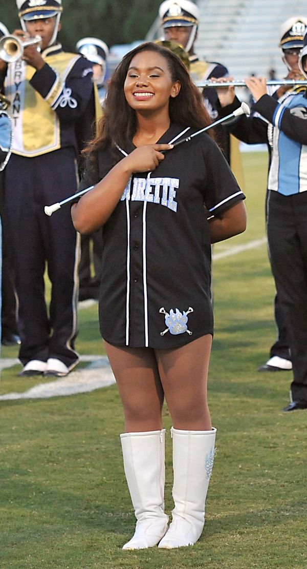 Majorette smiling confidently on the field with a baton, surrounded by band members.