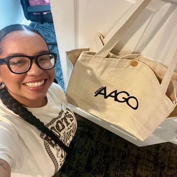 Smiling woman with glasses taking a selfie near a door with a beige AAGO tote bag.