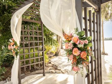 A decorated wedding arch with flowing white fabric and pink flowers outdoors.