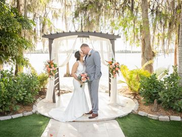 Bride and groom share a kiss under a floral arch by the water.