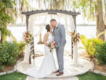 Bride and groom share a kiss under a floral arch by the water.