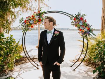 A groom standing in front of a floral arch by the water.
