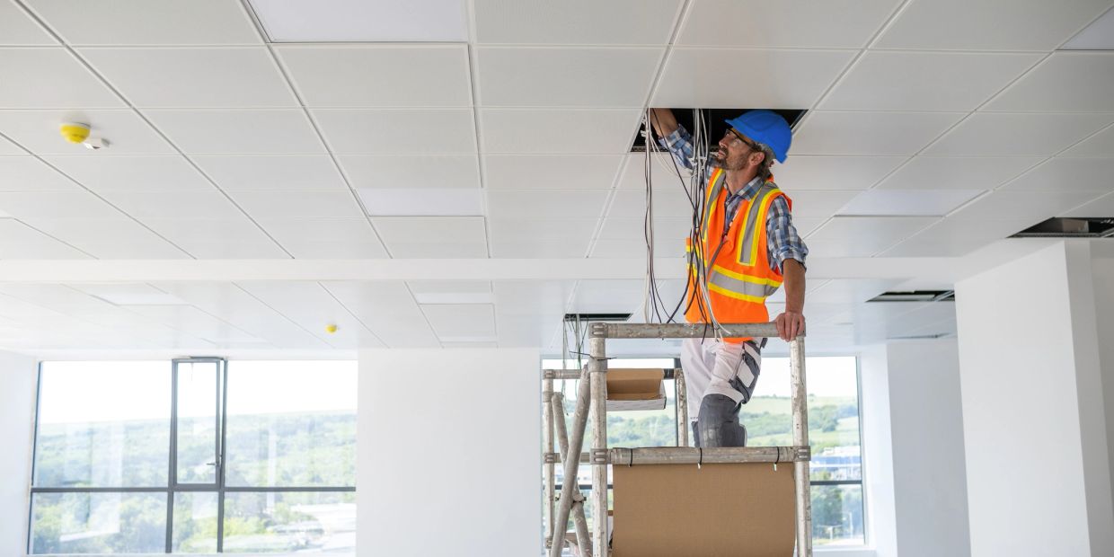 Electrician in safety gear working on ceiling wiring from scaffold.