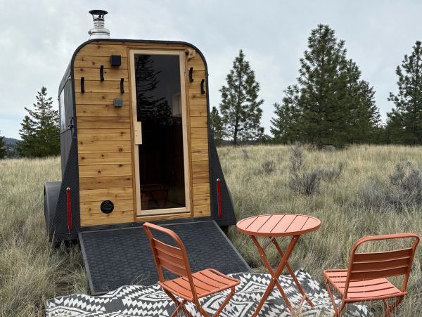 Compact wooden sauna trailer with outdoor seating in a natural setting.