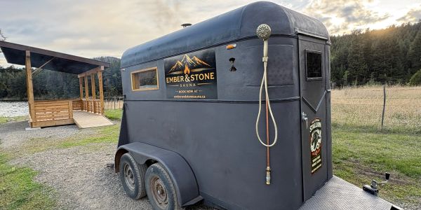A black trailer with Ember &amp; Stone Sauna signage parked outdoors near a wooden deck.