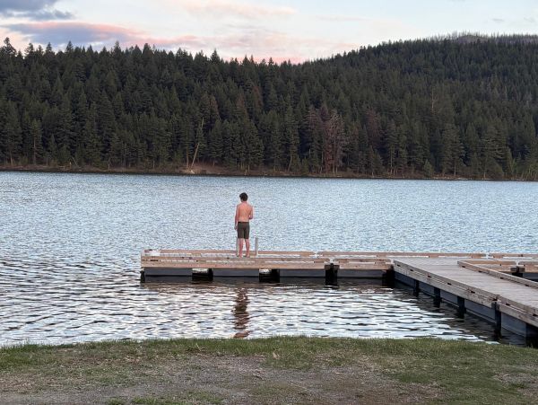 A boy stands alone on a wooden dock by a serene lake surrounded by forest.