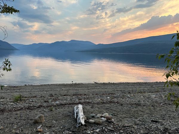 Calm lakeside at sunset with mountains and a fire pit on the shore.