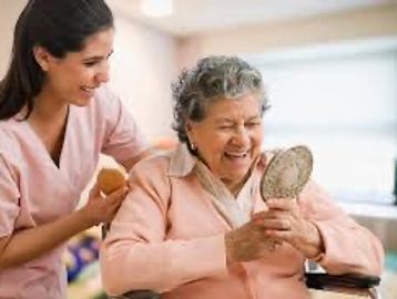 A caregiver helps an elderly woman smile while holding a mirror.
