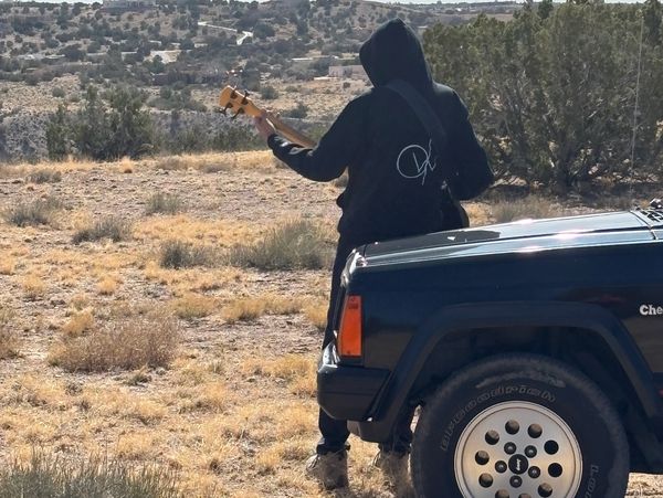 Person playing guitar near a black vehicle in a dry, open landscape.