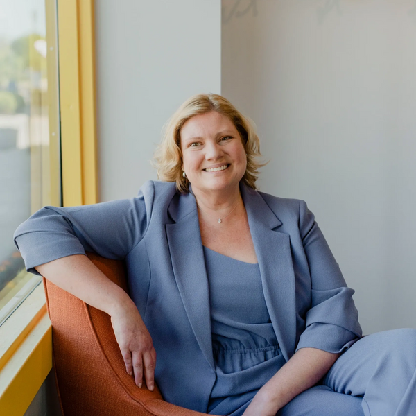 Smiling woman in a blue suit sitting comfortably on an orange chair near a window.