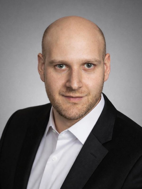 Professional headshot of a confident bald man in a black suit and white shirt.