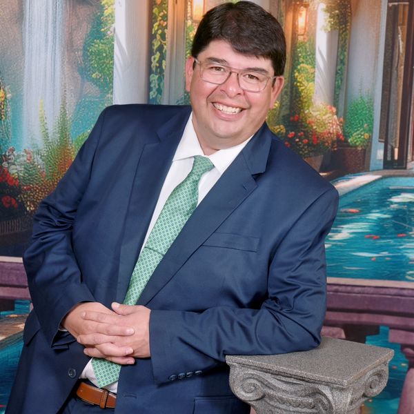 Man in blue suit smiling, leaning on a stone column with a scenic background.