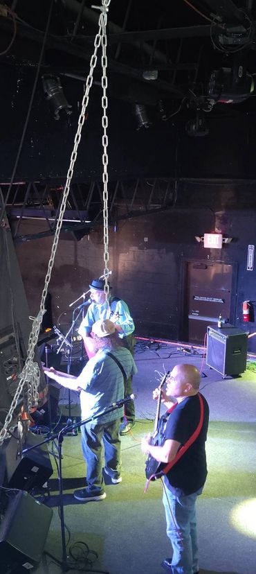 Three musicians perform on stage under stage lighting and hanging chains.