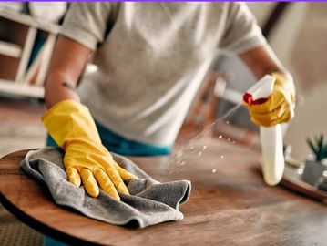 A white male holding a white spray bottle, wearing yellow rubber gloves cleaning a brown table.