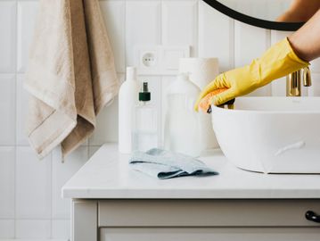 A white person wearing a yellow rubber glove, holding a sponge cleaning a mirror covered in soap.