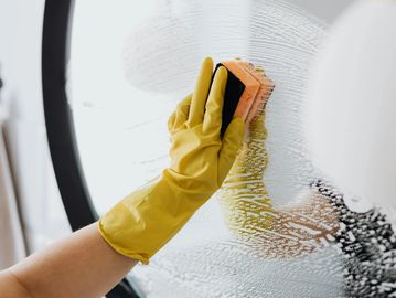 A white person wearing a yellow rubber glove, holding a sponge cleaning a mirror covered in soap.
