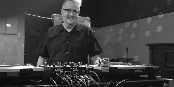 Smiling DJ standing behind a turntable setup in a warmly lit room.