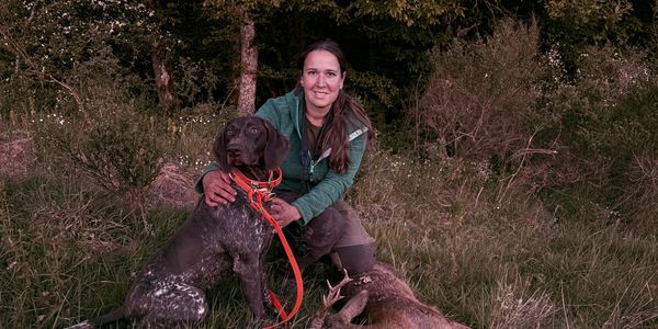 Frau kniet mit Hund und erlegtem Wild im Wald.
