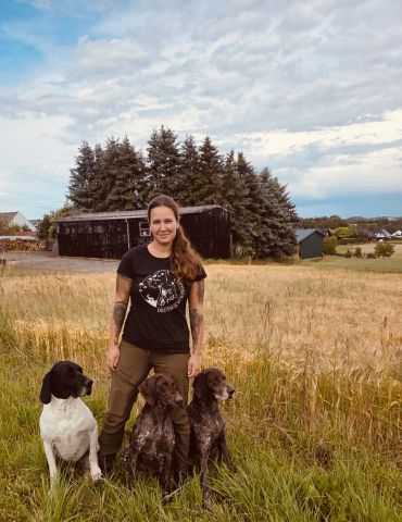 Woman standing with three dogs in a field under a cloudy sky.