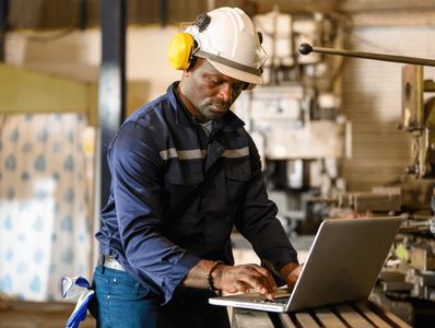 Industrial worker wearing safety gear using a laptop in a factory.