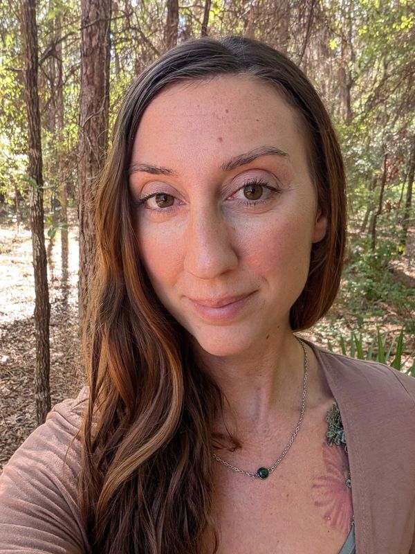 A woman with long brown hair stands in a sunlit forest, smiling gently.