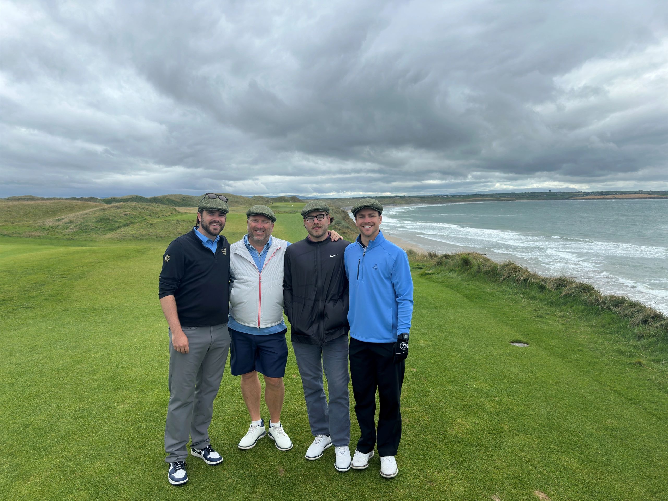 Four men in golf attire posing on a golf course by the sea.