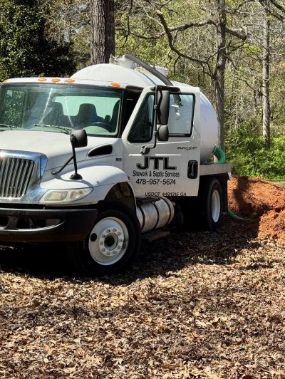 A white septic service truck parked in a wooded area with dry leaves.