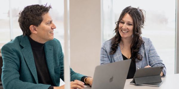 Two colleagues smiling and discussing work with laptops in a bright office.