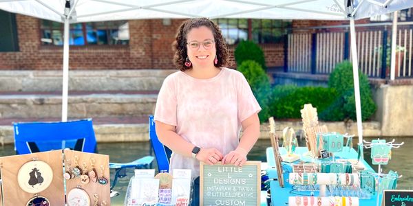 Woman smiling behind a craft booth at an outdoor market.