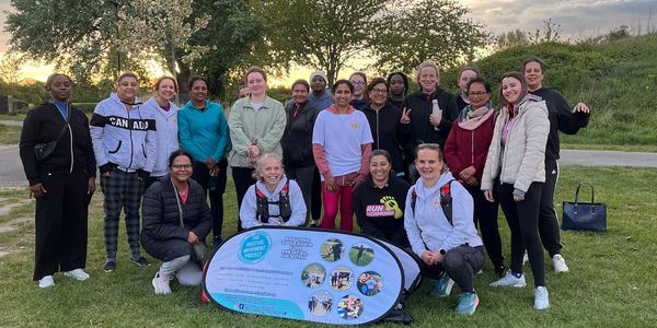 A diverse group of women posing outdoors with a community project banner.