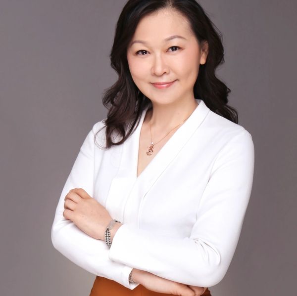 Confident woman in white blouse smiles with arms crossed against gray background.