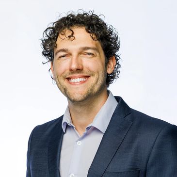 Smiling man with curly hair in a suit jacket against a white background.