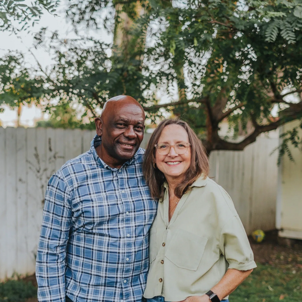 Smiling couple standing together outdoors with trees and a white fence in the background.